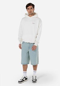 Fehér kapucnis pulóver elülső zsebbel és a mellkason logóval, világos farmer rövidnadrággal és fekete-fehér tornacipőkkel párosítva.