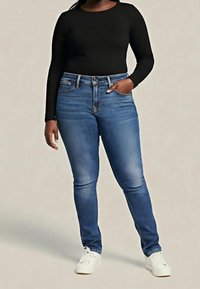 Personne portant un haut noir à manches longues, un jean bleu ajusté, des baskets blanches, debout avec une main dans la poche, devant un fond uni.