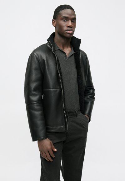 Schwarze Lederjacke mit Reißverschluss, hohem Kragen und Seitentaschen. Trägt man über einem dunkelgrauen Poloshirt, kombiniert mit dunkler Hose. Glatte Textur.