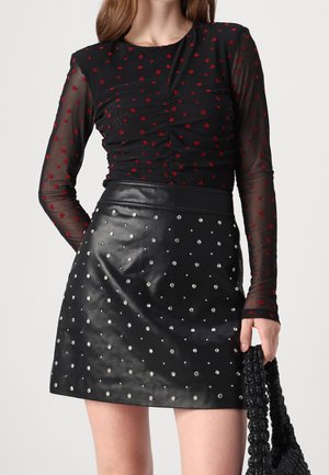 Haut en mesh noir à manches longues avec motifs de cœurs rouges associé à une mini-jupe en cuir noire ornée de clous argentés, tenant un sac noir texturé.