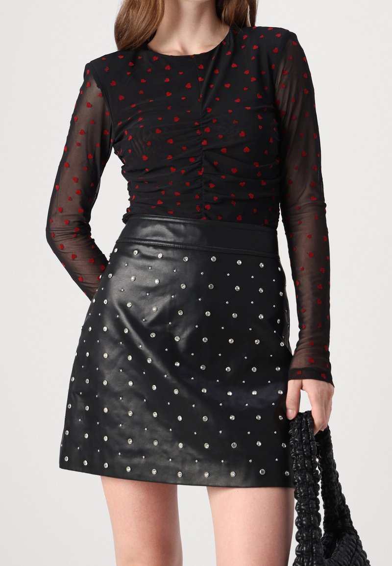 Haut en mesh noir à manches longues avec motifs de cœurs rouges associé à une mini-jupe en cuir noire ornée de clous argentés, tenant un sac noir texturé.