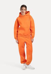 Conjunto de sudadera con capucha y pantalones de chándal en color naranja, fabricado en material suave, que cuenta con un bolsillo frontal, un corte relajado y puños acanalados. Zapatillas blancas.