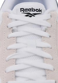 Weißer Sneaker mit einem beigen Wildlederpaneel, weißen Schnürsenkeln und einem schwarzen Reebok-Logo auf der Zunge. Das Material ist glatt und strukturiert.