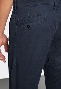 Marinblå bomullbyxor med en texturerad yta, med en bakficka som har en svart knapp och kontrasterande blå sömmar längs sidan.