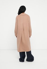 Cappotto lungo beige con colletto, spalle cadenti e una piega sul retro. Indossato sopra pantaloni scuri a gamba larga. Texture liscia.