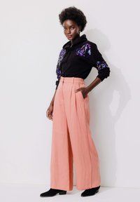Un sweat à capuche noir orné de sequins floraux violets, associé à un pantalon rose taille haute à jambe large. Des bottines noires à la cheville complètent le look.