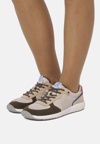 Beige Turnschuhe mit braunen Stoffakzenten, texturierten Schnürsenkeln, einem blauen Logoaufnäher und einer roten und blauen Fersenkappe. Gummisohle.