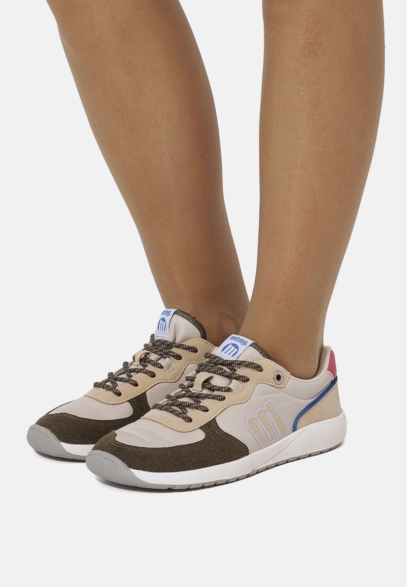 Beige Turnschuhe mit braunen Stoffakzenten, texturierten Schnürsenkeln, einem blauen Logoaufnäher und einer roten und blauen Fersenkappe. Gummisohle.
