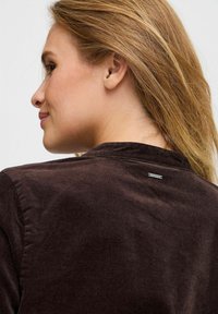 Brown corduroy jacket featuring a subtle texture, rounded collar, and a small metal tag at the back neckline.