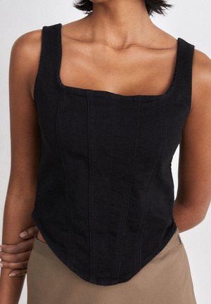 Kobieta nosząca czarny top bez rękawów z kwadratowym dekoltem i pionowymi detalami szwów, w połączeniu z jasnobrązowymi spodniami.