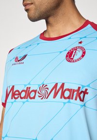 Vīrietis, kurš valkā gaiši zilu Feyenoord Rotterdam futbola kreklu ar sarkaniem akcentiem un MediaMarkt sponsora logotipu uz krūšu daļas.