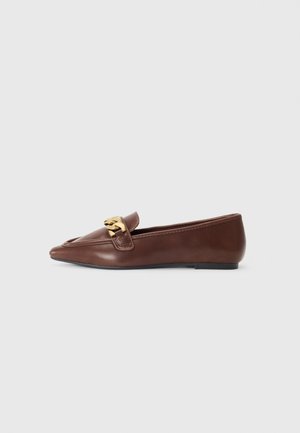 Bruine leren platte loafer met een gouden kettinkje bovenaan en een puntige neus op een lichtgrijze achtergrond.
