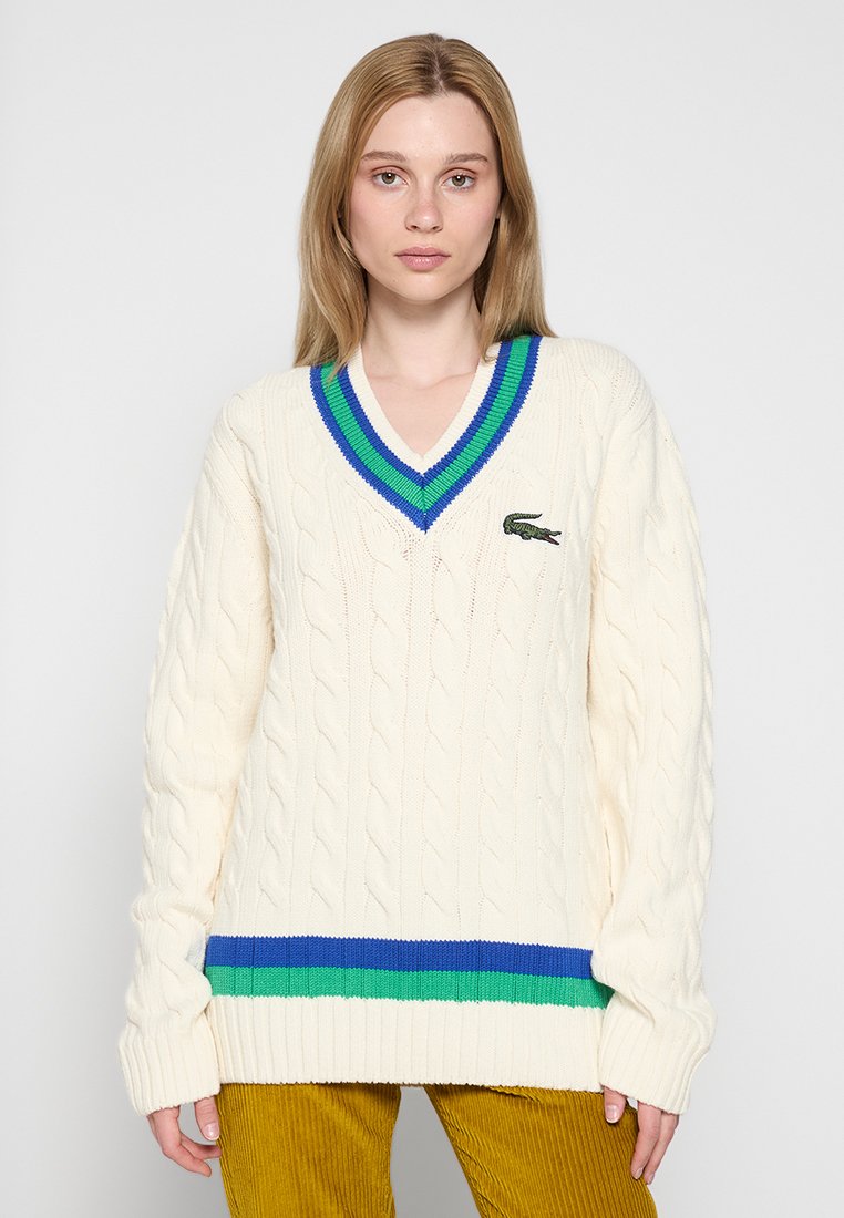 Jeune femme portant un pull en maille torsadée crème avec un col en V, des rayures bleu et vert, et un petit logo de crocodile sur la poitrine.