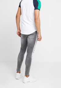 Jeans de mezclilla gris ajustados con rayas blancas laterales, usados con una camisa de color bloqueada en blanco y azul de manga corta, combinados con zapatillas blancas.