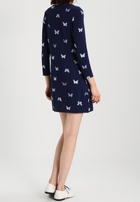 Mörkblå klänning i mjukt tyg med trekvartslånga ärmar, prydd med ett mönster av färgglada fjärilar. Den har en rund halsringning och en avslappnad passform.