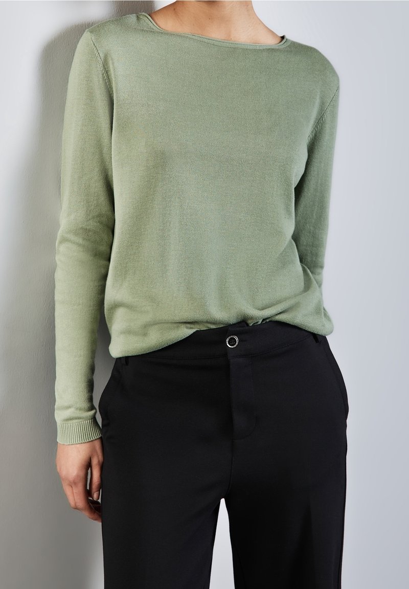Personne portant un pull vert clair à manches longues rentré dans un pantalon noir taille haute, debout contre un mur clair uni.