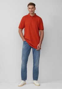 Rotes Poloshirt mit Kragen, kurzen Ärmeln und einem dezenten Logo, kombiniert mit hellblauen Jeans und weißen Turnschuhen. Glatte Textur insgesamt.