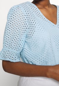 Jasnoniebieska bluzka z krótkim rękawem i dekoltem w szpic, z haftowanymi otworami i falbankami na końcach rękawów. Teksturowana tkanina i luźny krój.