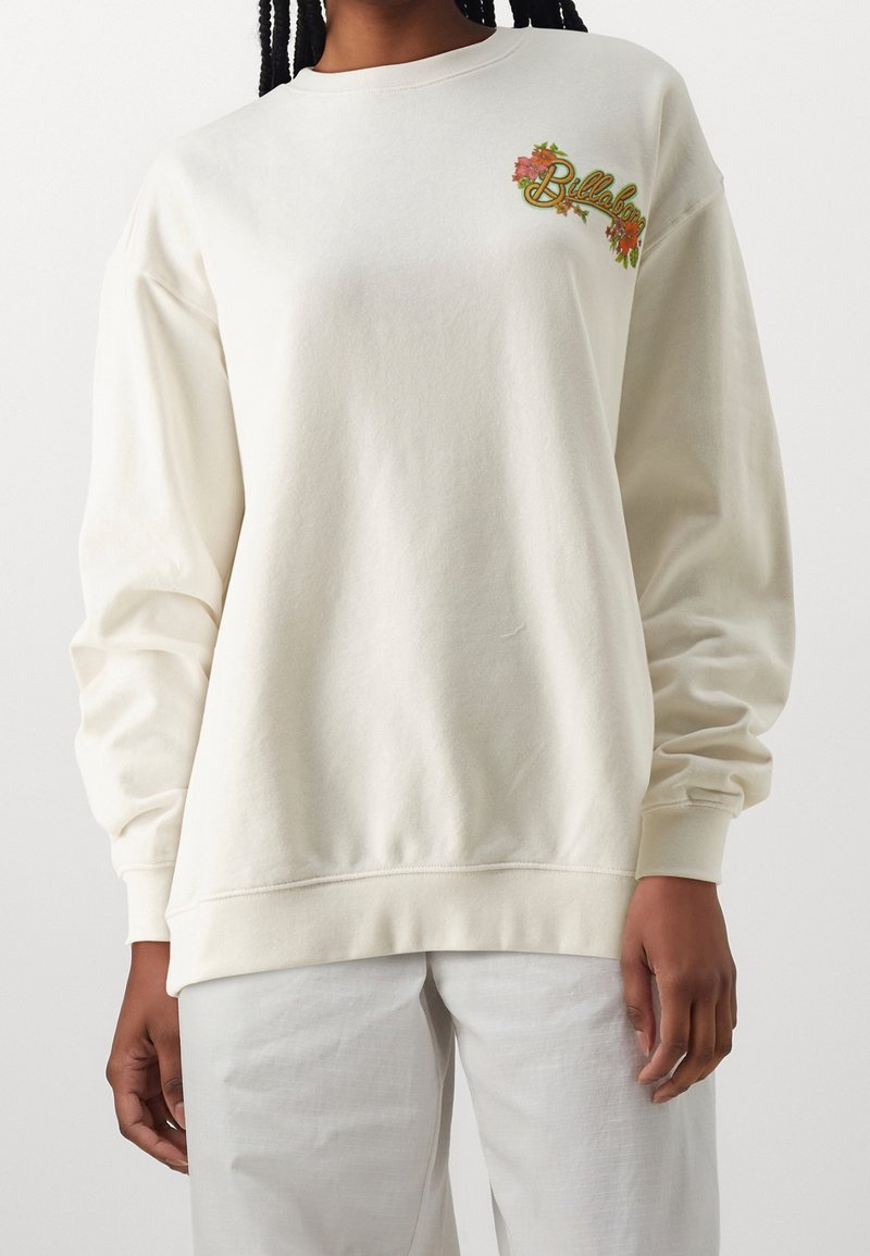 Personne portant un sweat-shirt à manches longues blanc cassé avec un petit motif floral coloré et un texte sur la partie supérieure gauche de la poitrine.