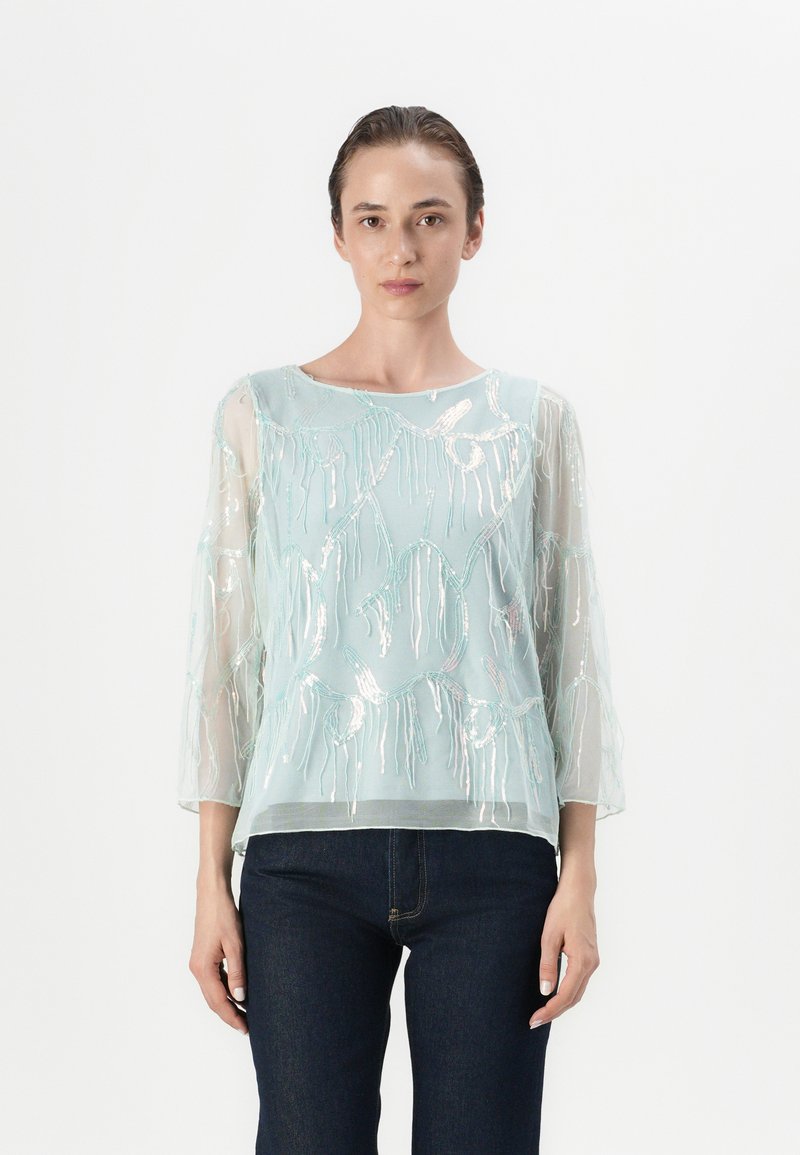 Blusa leggera azzurro chiaro con intricate ricami in argento e maniche a tre quarti, presenta un colletto rotondo e un design fluido.