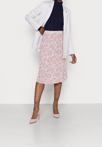 Kvinne iført en marineblå genser med høy hals, lett lilla blazer, mønstrete midi-skirt og rosa høye hæler, står mot en enkel hvit bakgrunn.