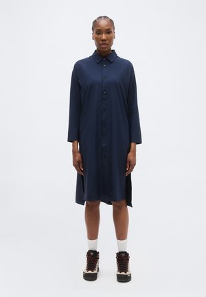 Vrouw met gevlochten haar, gekleed in een marineblauwe overhemdjurk met knopen, witte sokken en beige wandelschoenen, staand tegen een effen witte achtergrond.