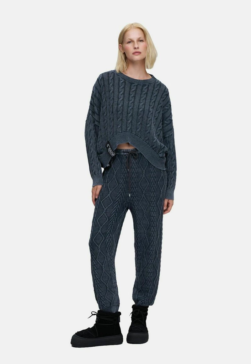 Femme portant un pull en maille torsadée gris foncé et un pantalon de jogging assorti avec des bottes d'hiver noires à lacets, debout devant un fond uni.