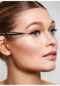 Flüssiger Eyeliner-Stift, der nah am Auge gehalten wird; das Model hat glatte Haut, dezentes Make-up und eine neutrale Lippenfarbe; blaue Augen und braune Haare.
