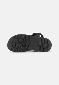 Svart sandal med en texturerad gummisula och remdesign med en logotyp präglad på undersidan. Klacken erbjuder grepp med spår.