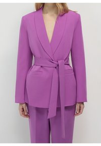 Fioletowa marynarka z głębokim dekoltem w kształcie litery V, szerokimi klapami i związanym paskiem w talii. Dwie przednie kieszenie oraz gładki, usztywniony materiał.