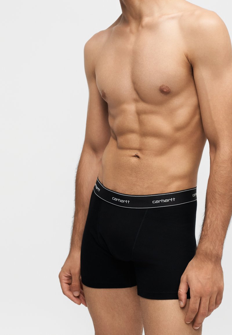 Athletischer männlicher Oberkörper in schwarzen Carhartt-Boxershorts, stehend vor einem schlichten hellen Hintergrund mit entspannt seitlich hängenden Armen.
