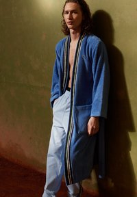 Accappatoio in terry di colore blu con cappuccio e tasche, caratterizzato da righe nere, verdi e bianche lungo i bordi; indossato sopra pantaloni della tuta blu chiaro.