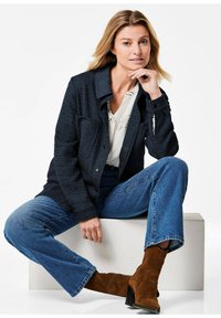 Blue textured jacket with two front pockets, paired with a white blouse and flared blue jeans. The model wears brown suede ankle boots.