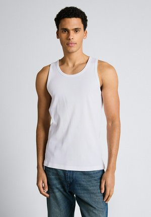 Młody mężczyzna ubrany w białą koszulkę na ramiączkach i niebieskie dżinsy stoi na tle jednolitego jasnoszarego tła.