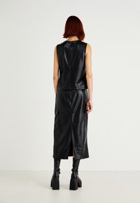 Schwarzes Leder-Midikleid mit ärmellosem Design, Reißverschluss auf der Rückseite und seitlichen Taschen. Kombiniert mit kniehohen Blockabsatzstiefeln.