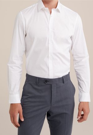 Mann trägt ein eng anliegendes weißes Hemd, in graue Anzughose gesteckt, steht mit entspannten Armen an den Seiten.