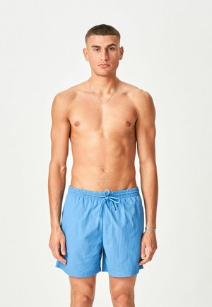 Young man with short hair stands facing forward, shirtless, wearing light blue swim shorts with a drawstring.
