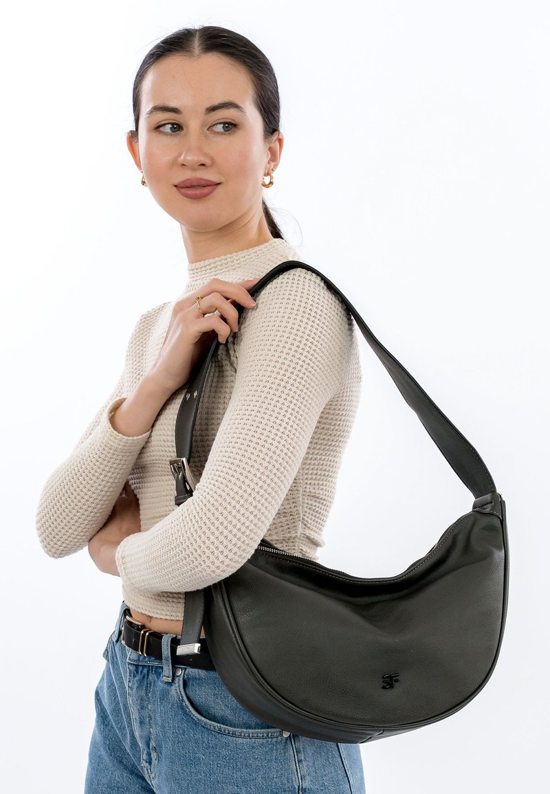 Woman in beige knit top and blue jeans carrying a large black leather shoulder bag with a wide strap, looking to the side.