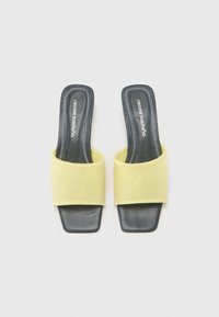 Sárga anyagból készült csúszó szandál, széles pánttal és fekete négyzet formájú orral. Lakkozott talppal rendelkezik, és varrott részletek díszítik a szélein.