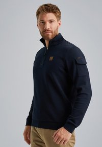 Sudadera de media cremallera en azul marino con un acabado texturizado, que presenta un cuello con cremallera y un bolsillo con parche en la manga izquierda.