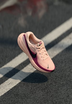 Nike running shoe in light pink with purple accents. Features a breathable mesh upper and a textured sole. Floating above a dark surface.