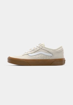 Zapatilla de ante beis claro con acentos de cuero blanco, puntera redonda, parte frontal con cordones y suela de goma marrón con superficie texturizada. Detalles de cuero perforado en la parte superior.