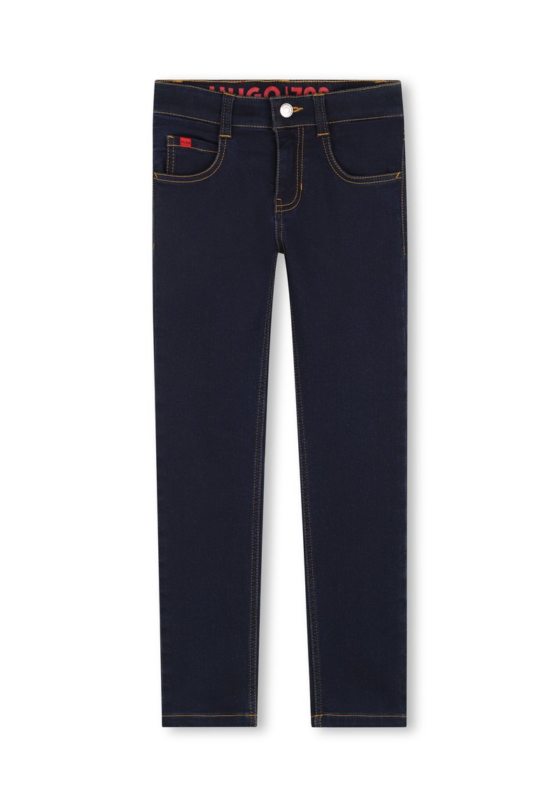 Jean bleu foncé avec une coupe slim, présentant des coutures jaunes, cinq poches et un petit logo rouge sur le devant. Texture denim lisse.