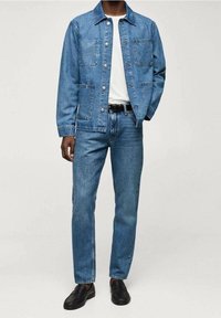 Veste en denim avec deux poches poitrine, accompagnée d'un jean droit en denim. Les deux sont bleu moyen, en tissu texturé, portés avec des mocassins noirs.