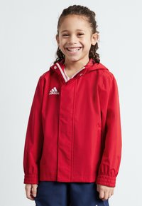Röd vattentät jacka med huva, elastiska ärmslut och dragkedja fram. Har en vit Adidas-logotyp på bröstet och en slät textur.