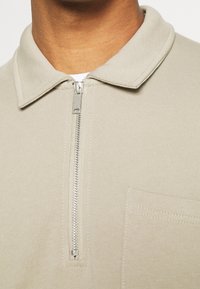 Chemise beige clair à fermeture éclair avec un col pointu, une texture côtelée douce, et une poche poitrine gauche. La fermeture éclair est en métal.