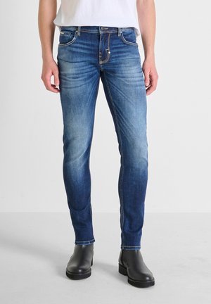 Vaqueros de mezclilla azul desgastada con un ajuste slim, con costuras en contraste, cinco bolsillos y cierre con cremallera, combinados con botas negras de tobillo.