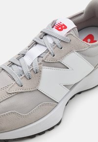 Sneaker grigio chiaro realizzato con una combinazione di materiali in tessuto e suede. Accenti bianchi e un logo rosso sulla linguetta. Lacci tondi.