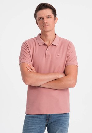 Helles rosa Polohemd mit Kragen und kurzen Ärmeln, versehen mit einem kleinen gestickten Logo auf der linken Brust. Getragen mit blauer Denim-Jeans.