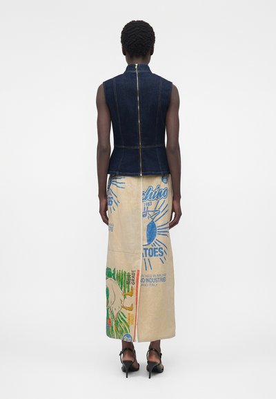 Άτομο που φοράει αμάνικο σκούρο τοπ από τζιν με φερμουάρ στην πλάτη και μακριά μπεζ φούστα με πολύχρωμα τυπωμένα σχέδια, στέκεται με την πλάτη στην κάμερα.